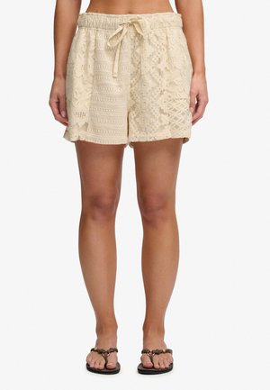 Frau trägt beige Shorts mit Spitzenmuster und Kordelzug an der Taille sowie schwarze geflochtene Sandalen; steht vor weißem Hintergrund.