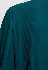 Tissu plissé vert sarcelle sur un vêtement ample couvrant une épaule et le haut du bras, montrant des plis fins verticaux et une texture.