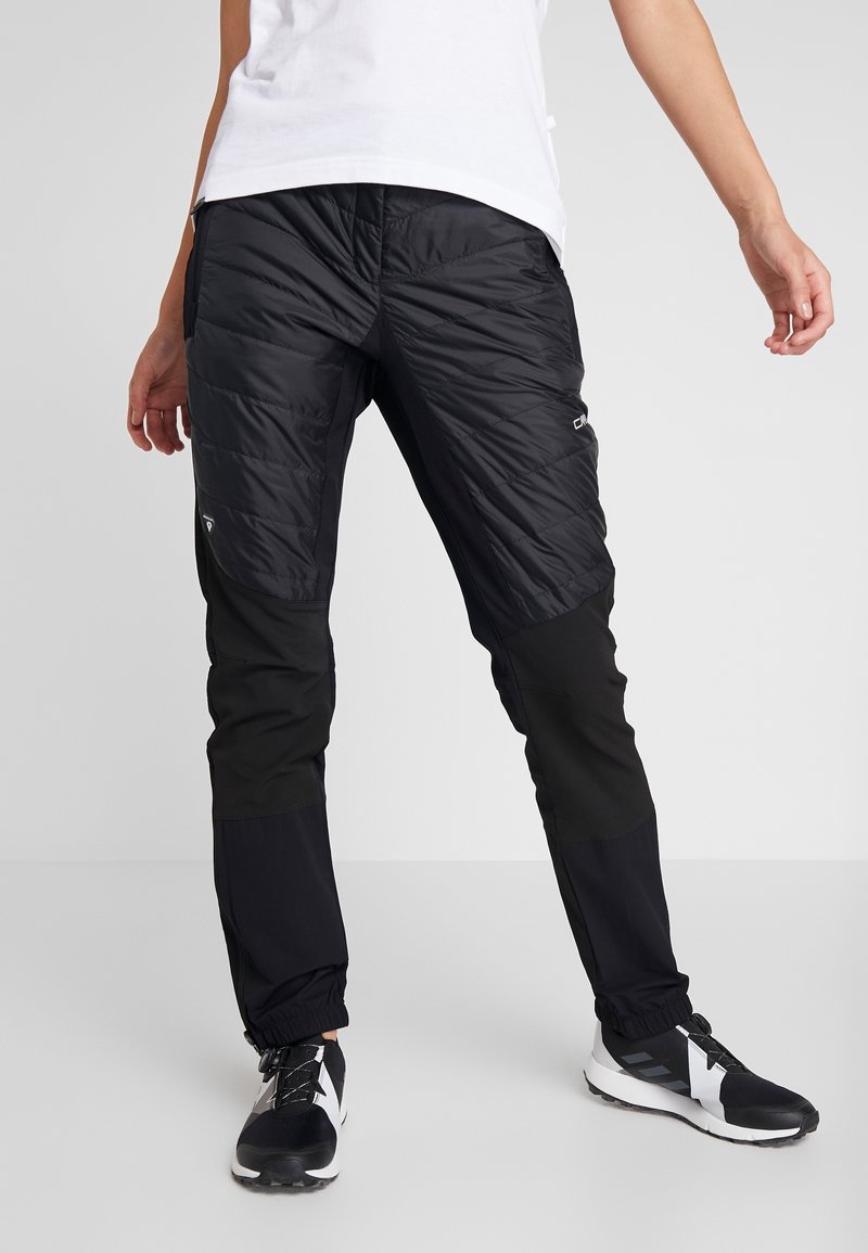 Personne portant un pantalon isolant noir avec la partie supérieure des cuisses matelassée et des chaussures de sport noires, debout devant un fond gris.