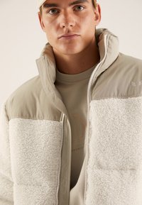 Beige zip-up jacket with a fluffy white lower section, smooth upper fabric, and a high collar; features a logo on the shoulder.