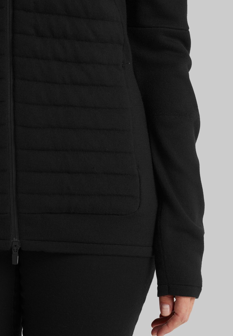 Nahaufnahme einer Person, die eine schwarze, gesteppten Jacke mit Reißverschluss trägt. Zu sehen ist der untere Oberkörper und der rechte Arm vor einem grauen Hintergrund.