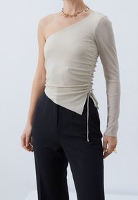 Beige one-shoulder top with ribbed texture, ruched detail on side, and drawstring at the waist, paired with black tailored pants.