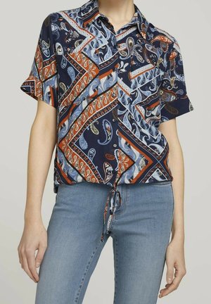 Kortærmet skjorte med knapper med blå, orange og hvide paisley- og geometriske mønstre, bundet i taljen, båret med lyseblå jeans.