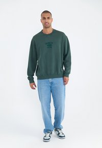 Donkergroene sweatshirt met de opdruk "BROOKLYN 1993 NEW YORK", gecombineerd met lichtblauwe jeans en zwart/witte sneakers. Glad oppervlak, casual ontwerp.