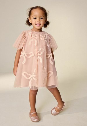 Ung pige iført en lyserød kjole med sløjfemønstre og glitrende lyserøde Mary Jane-sko, stående mod en neutral baggrund.