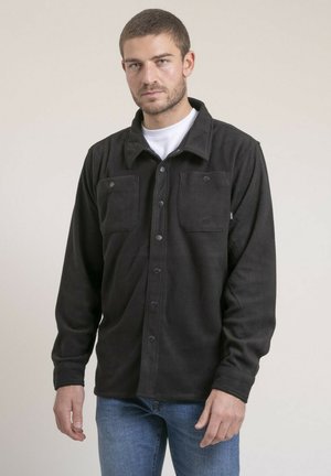 Homme portant une veste polaire noire à boutons sur une chemise blanche et un jean bleu, debout devant un fond beige uni.
