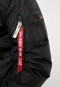 Chaqueta de nylon negra con un bolsillo con cremallera, un parche tejido y una etiqueta de tela roja que dice "RETIRAR ANTES DEL VUELO". Detalle de superficie texturizada.
