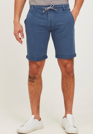 Blå denimshorts med opkrøllede manchetter, der har en snørebånds talje. Bæres med afslappede hvide sneakers og en tatovering synlig på benet.