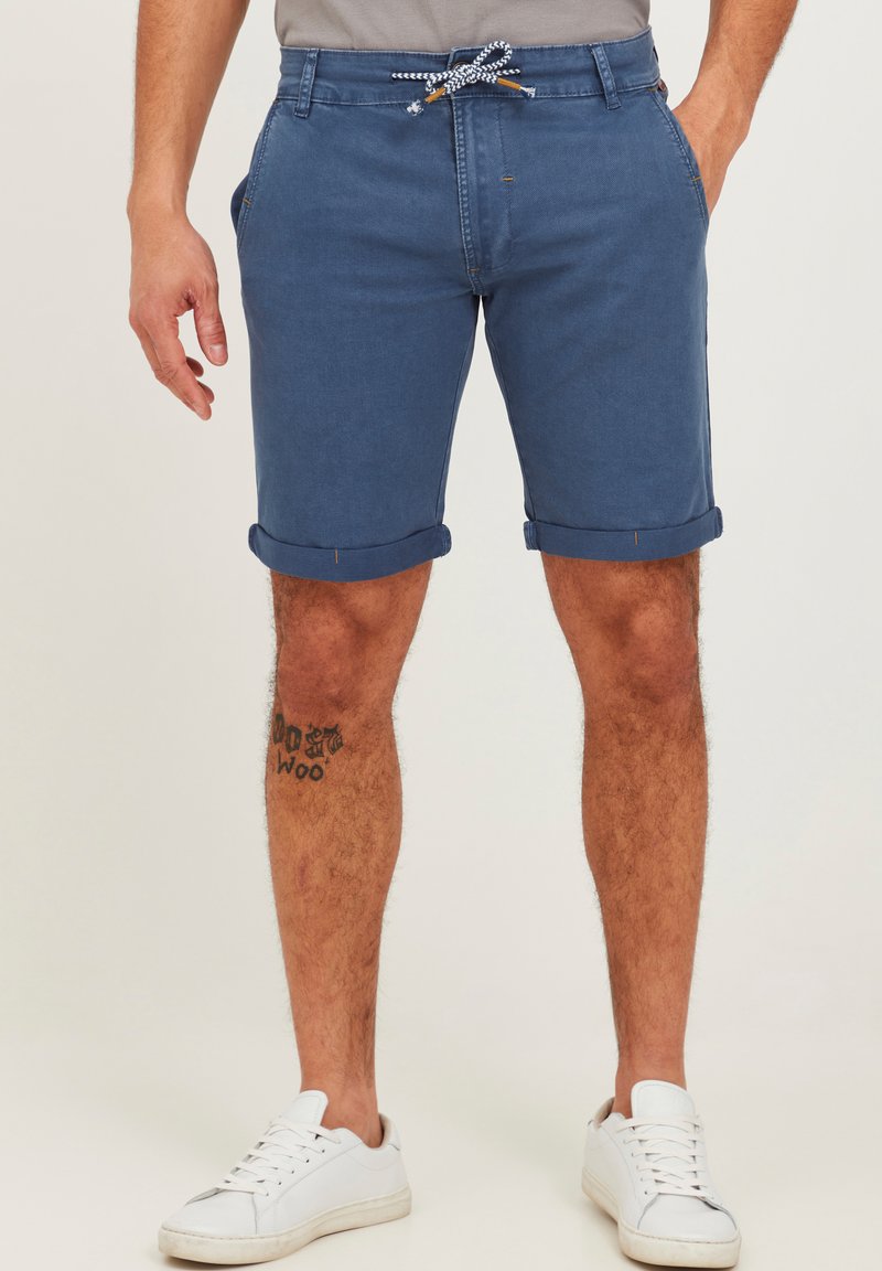 Sinine denim lühike püksid keeratud servadega, millel on paelaveoga vöö. Kandmine koos juhuslike valgete tossudega ja jala peal nähtav tätoveering.