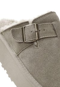 Grijze suède slof met een pluizige voering, voorzien van een metalen gesp en ton-sur-ton stiksels. De zool is uitgevoerd met een geribbeld patroon voor grip.