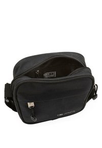 Bolso de hombro de tela negra con cierre de cremallera, correa ajustable y bolsillo interior. Presenta un logo de la marca en la parte frontal.