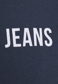Texto blanco "JEANS" impreso sobre un fondo de tela de canalé azul oscuro.