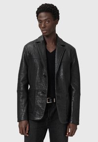 Schwarze Lederblazer mit strukturierter Oberfläche, geknöpften Revers, vorderen Knöpfen und zwei vorderen Taschen. Getragen über einem schwarzen Pullover.