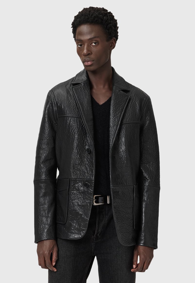 Schwarze Lederblazer mit strukturierter Oberfläche, geknöpften Revers, vorderen Knöpfen und zwei vorderen Taschen. Getragen über einem schwarzen Pullover.