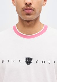 Κοντινό ενός ατόμου που φοράει λευκό πουκάμισο με ροζ γιακά και το κείμενο «NIKE GOLF» καθώς και ένα μαύρο λογότυπο με θέμα το γκολφ στο στήθος.