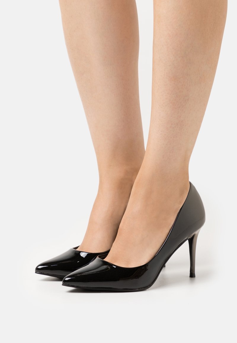 Jambes portant des escarpins noirs brillants à bout pointu avec des talons aiguilles fins sur fond blanc.