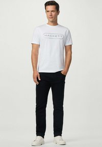Camiseta de cuello redondo blanca con el logo "HACKETT" en gris, combinada con jeans negros y zapatillas blancas. Diseño sencillo, fabricada en algodón.