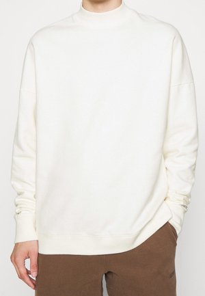 Personne portant un pull blanc uni à manches longues avec poignets et col côtelés, associé à un pantalon marron, les mains détendues le long du corps.