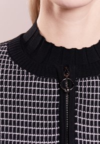 Black and white patterned zip-up sweater featuring a ribbed collar, textured fabric, and a circular zipper pull.