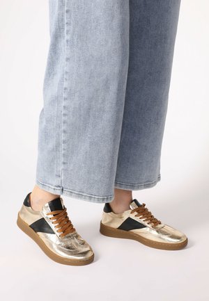 Gold metallic sneakers with black accents, featuring brown laces and a textured surface, set against light blue denim pants.