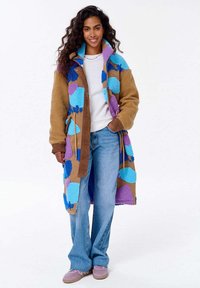 Uśmiechnięta kobieta z długimi kręconymi włosami, ubrana w kolorowy p�łaszcz z patchworku, biały top, niebieskie dżinsy i różowe trampki, stojąca z jedną ręką w kieszeni.