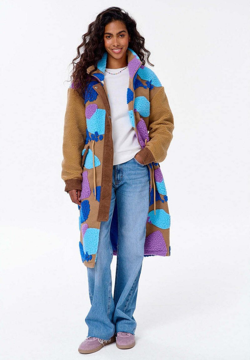 Uśmiechnięta kobieta z długimi kręconymi włosami, ubrana w kolorowy płaszcz z patchworku, biały top, niebieskie dżinsy i różowe trampki, stojąca z jedną ręką w kieszeni.