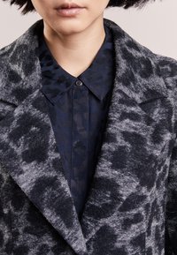 Wollenblend jas met een grijs en zwarte luipaardprint, voorzien van een geknoopte kraag en een marineblauwe blouse eronder. Gelaagde stof.