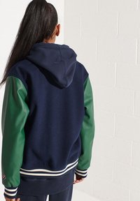 Vrouw met lang haar in een paardenstaart, gekleed in een donkerblauwe hoodie met groene mouwen en gestreepte manchetten, staand tegen een witte bakstenen muur.