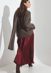 Brauner Strickpullover mit verlängertem linken Ärmel, kombiniert mit einem fließenden burgunderfarbenen Satinkleid. Modell trägt hohe Stiefel. Schatten sichtbar.