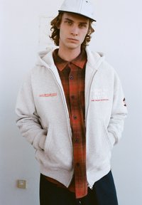 Šedá mikina na zip s kapucí se stahovací šňůrkou, s červenými grafikami. Nositelná přes červenou a černou károvanou košili na knoflíky s černými knoflíky.