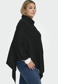 Poncho en tricot noir avec un col haut et une ouverture à l'avant. Présente une finition texturée et un ourlet asymétrique, associé à un jean bleu.