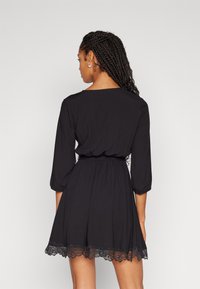 Robe noire avec des manches trois-quarts et une taille cintrée, présentant une finition en dentelle à l'ourlet. Le tissu lisse ajoute douceur et fluidité.