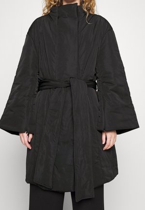 Vrouw die een zwarte gewatteerde jas draagt met brede mouwen, een hoge kraag en een stoffen ceintuur die in de taille is gestrikt.