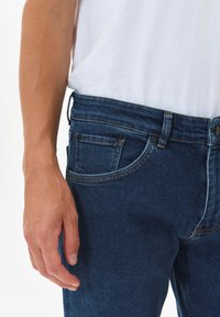 Temno sijoči jeans z oprijetim krojem, z vidnimi šivi, sprednjim žepom in gumbom za zapiranje. Tkanina ima gladko teksturo.
