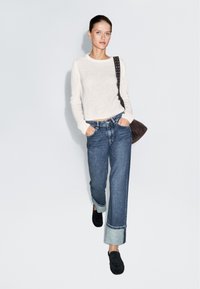 Jasnobeżowy dziany sweter, niebieskie dżinsy z wysokim stanem i podwiniętymi nogawkami oraz czarne wsuwane buty; modelka nosi ciemnobrązową torbę na ramię.