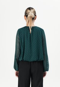 Woman with blonde hair in a ponytail wearing a dark green zigzag patterned blouse with sheer sleeves and black pants, facing away.