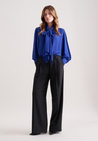 Blusa blu con un grande fiocco al collo, abbinata a pantaloni a gamba larga a righe nere e tacchi a punta neri. Tessuto dalla texture liscia.
