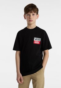 Camiseta de algodón negra con mangas cortas, que presenta un logo de bolsillo frontal en blanco y rojo. El ajuste es holgado. Se lleva con pantalones beiges.