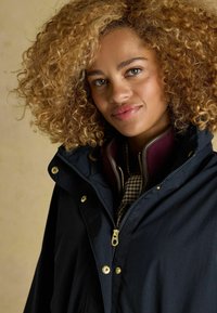 Jachetă impermeabilă de marină cu accesorii aurii, având un guler înalt, închidere cu fermoar și un model în carouri dedesubt. Material texturat.