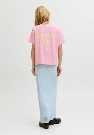 Femeie cu păr blond purtând un tricou roz "Aperitivo Club", fustă din dantelă albastru deschis și pantofi negri, stând cu spatele pe un fundal alb.