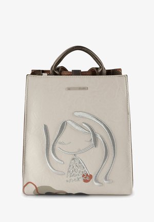 Cremefarbene Handtasche mit metallisch-silbernem, besticktem, abstraktem Gesicht und braunen, strukturierten Griffen, mit einer kleinen Logo-Plakette oben in der Mitte.