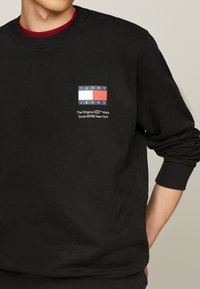 Sudadera negra con un logo rectangular en rojo, blanco y azul. El texto incluye "TOMMY JEANS" y "La Marca Original Desde 1985 Nueva York."