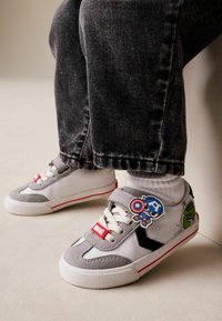 Zapatillas grises y blancas con detalles en rojo, con una tira de velcro y parches de personajes de Marvel, sobre pantalones de mezclilla oscuros.