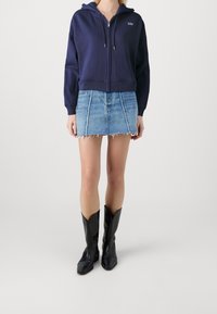 Sudadera azul marino con cremallera frontal y logotipo, combinada con una minifalda de denim con bordes deshilachados y un corte ceñido. Botas de vaquero negras completan el conjunto.