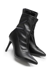 Pár černých kožených kotníkových bot s špičatou špičkou, bočním zipem a vysokými štíhlými podpatky na bílém pozadí.