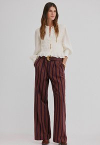 Biała, teksturowana bluzka z falbaniastym dołem, połączona z bordowymi, paskowanymi spodniami o szerokich nogawkach, podkreślona brązowym paskiem i złotymi akcentami.