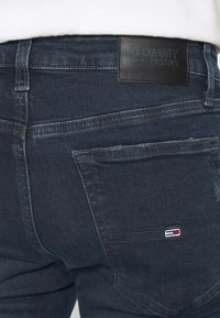 Tummansiniset farkut, joissa on nahkainen Tommy Jeans -tunnus, ommellut yksityiskohdat ja pieni brodeerattu logo takataskussa.