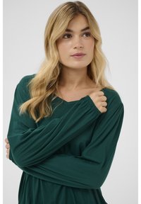 Grünes Langarmshirt mit V-Ausschnitt aus weichem Stoff. Mit gerafften Ärmeln und lockerem Schnitt, zeigt eine glatte Textur und schlichtes Design.