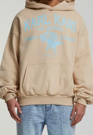 Person trägt einen beigen Karl Kani Hoodie mit blauem Schriftzug und Adlergrafik, kombiniert mit hellblauen Jeans, Hände locker an den Seiten.