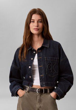 Jeune femme aux longs cheveux bruns portant une veste en jean foncée par-dessus un top court blanc et un pantalon beige taille haute, les mains dans les poches.